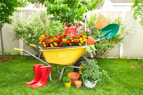 A garden scene featuring a bright yellow wheelbarrow filled with vibrant red and yellow flowers set on a well-maintained lawn. Behind the wheelbarrow, there is a white fence with lush green foliage and trees providing shade. To the left, a pair of red rubber gardening boots rests on the grass, while nearby, several small terracotta and plastic pots are arranged around a potted plant with broad green leaves. A red watering can and an orange leaf rake are placed next to the wheelbarrow, alongside a green-handled shovel and a large orange broom. The background shows a tidy outdoor space suited for gardening and lawn care, exemplifying a typical landscaped garden area in Crouch End with natural lighting and a calm, outdoor environment. Lawn Mowing Crouch Endâ€™s gardening service specializes in maintaining such vibrant, well-kept garden spaces in the area, evident through the neatly arranged plantings and clean, organized tools.