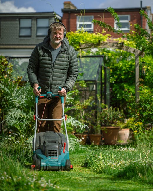 A man with light skin, short brown hair, and a beard operates a blue and black lawn mower, cutting a well-maintained grassy lawn in a private garden. He is dressed in a dark puffer jacket and brown tr